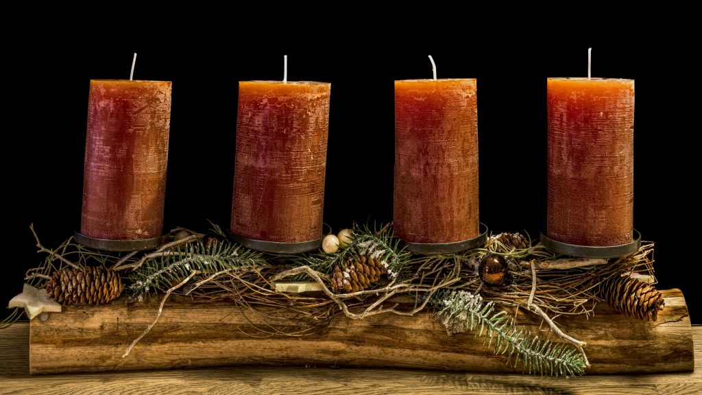 Four rustic advent candles on a wooden display with pine cones and greenery.